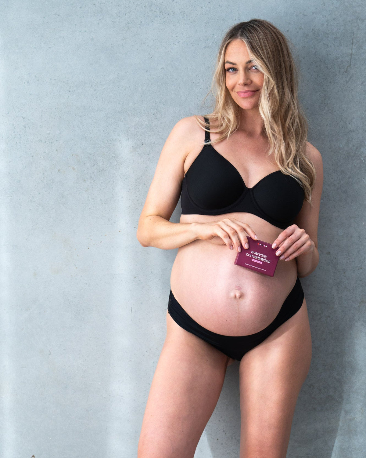 Pregnant woman wearing black maternity nursing bra and matching briefs, holding Hotmilk x Everyday Conversations Self Love Edition card pack against her bump